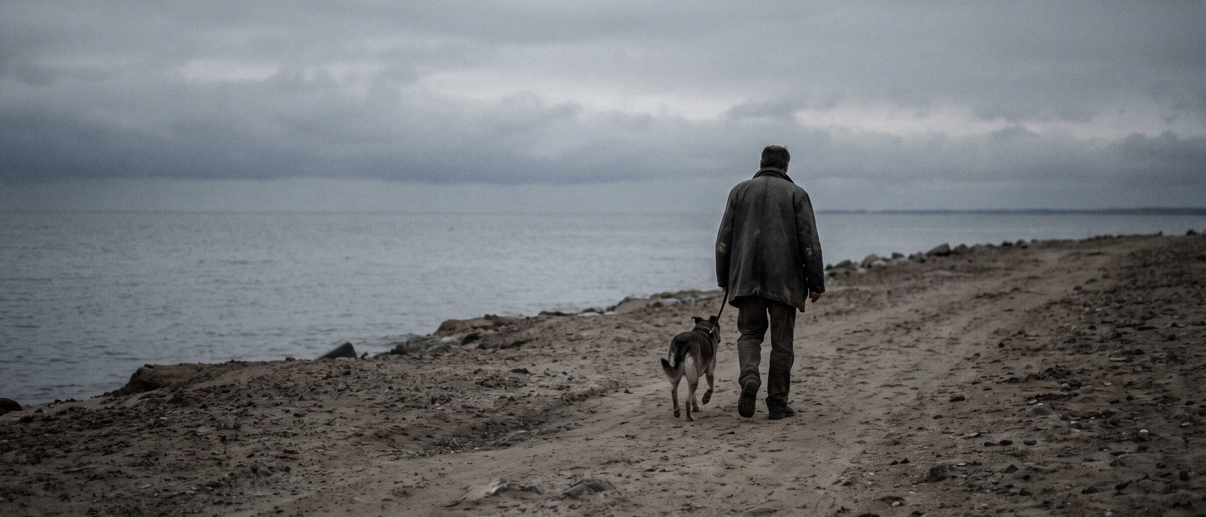 A man walks his dog along an empty shoreline under an overcast sky
